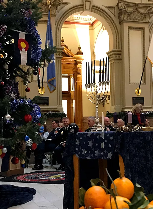A holiday Menorah with eight unlit candles on a table next to the Capitol's holiday tree. People are seated in chairs for the ceremony.